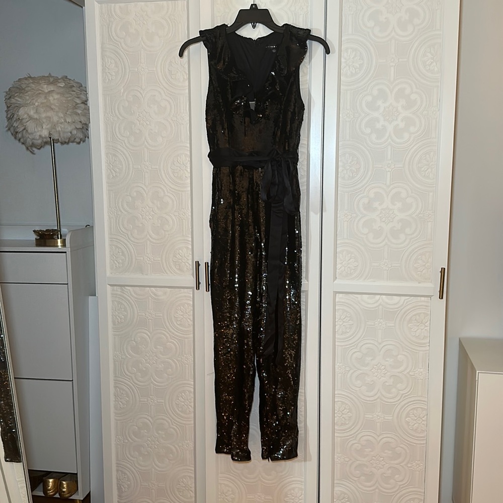 Black sequin jumpsuit with ruffle neckline and satin sash. Tapered pant leg